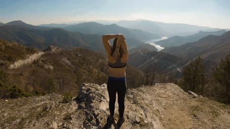 una atleta estirándose en la cima de una montaña con un hermoso lago en el cañón frente a ella.