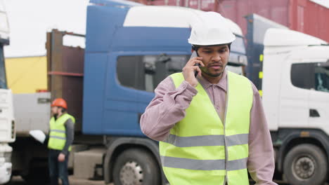 worker wearing vest and safety helmet organizing a truck fleet in a logistics park while talking on the phone 1