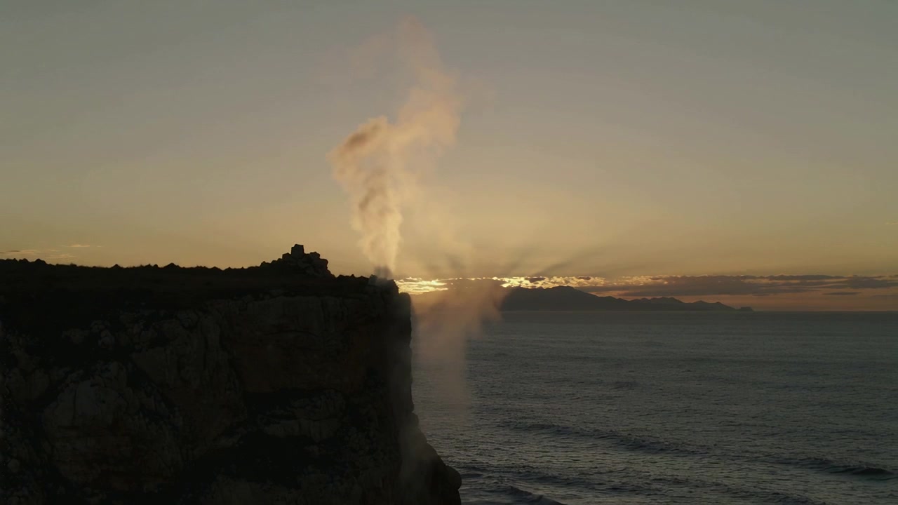 Spray de água subindo da borda de um penhasco rochoso ao pôr do sol, impulsionado pelo impacto das ondas e pelo vento