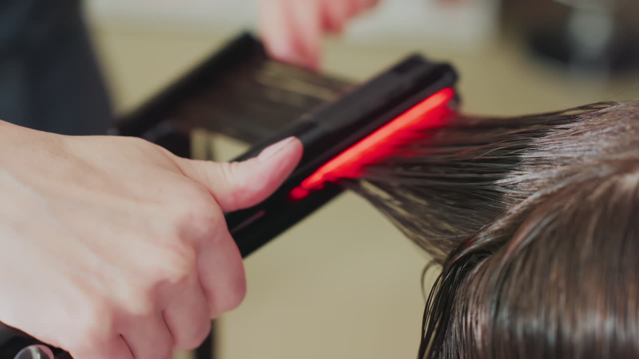 Vådt hår i salon, stylist bruger infrarødt glattejern til at glatte langt kaukasisk hår, nærbillede af opvarmede plader, der glider gennem hårlokkerne, klinisk professionel stemning og blankt resultat