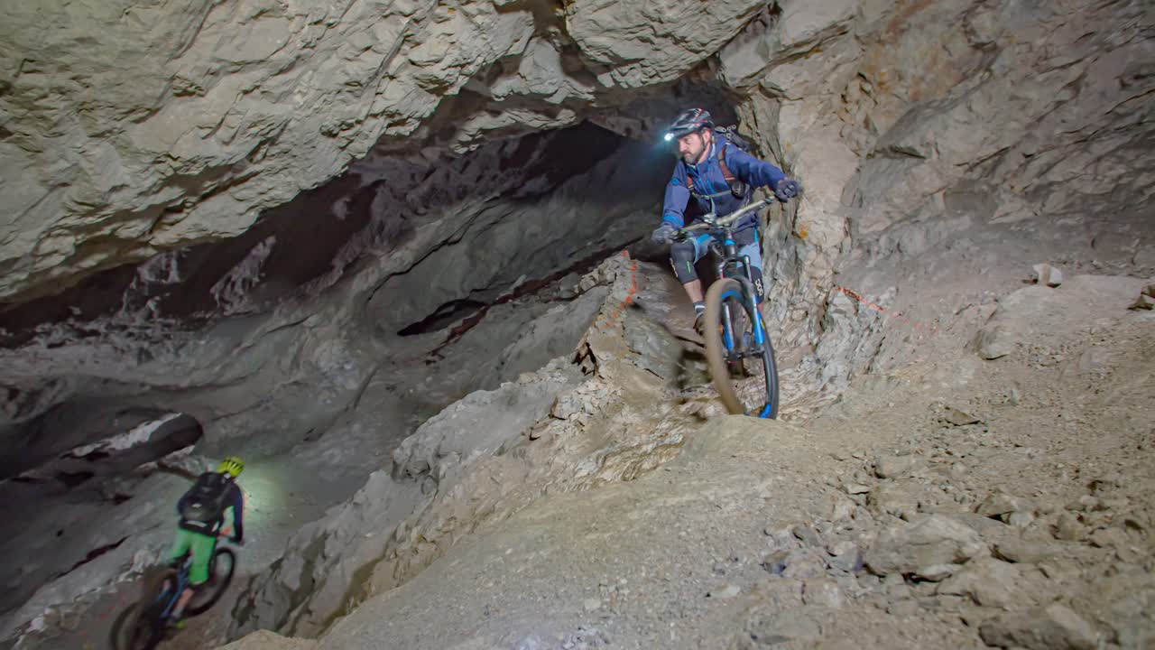 Two sporty cyclists on professional mountain bikes cross a dark mine with rocky narrow walls on a underground tour