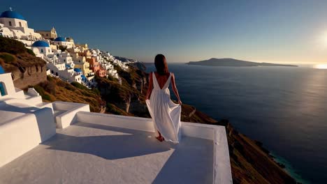 A woman in a white dress stands on a rooftop overlooking a coastal village at sunset