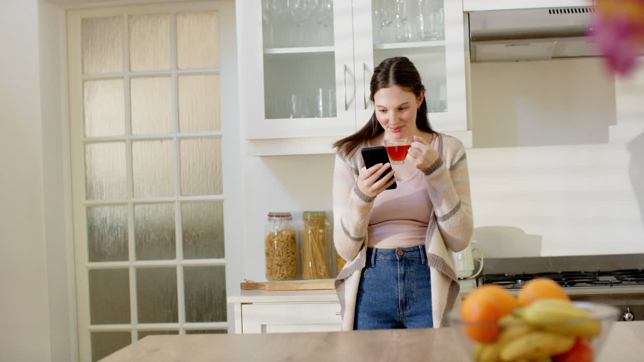 mujer caucásica feliz usando teléfono inteligente y bebiendo té en la cocina en casa, espacio de copia, cámara lenta