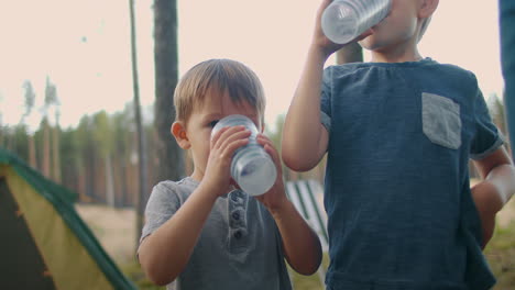 two little boys are drinking water brothers are resting in campsite with parents family travelling at nature