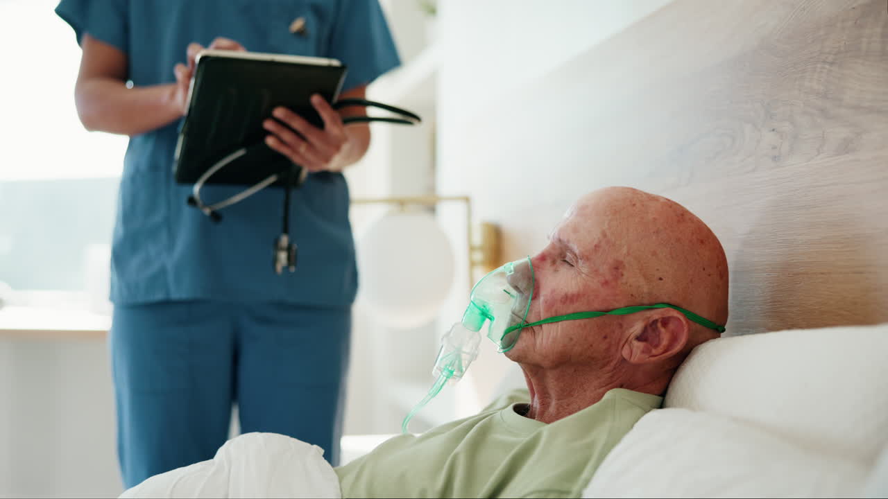 Elderly man receiving medical care in a hospital bed with an oxygen mask while a nurse checks his status on a tablet