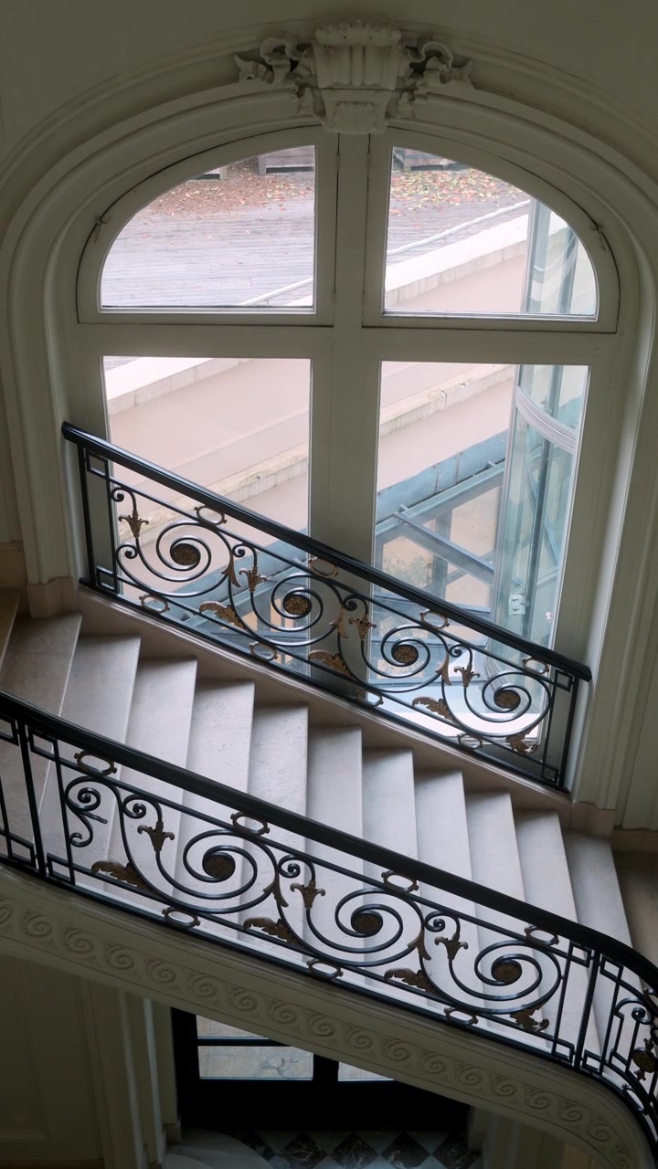 Pemandangan interior tangga Paris yang elegan dengan pagar besi berukir dan jendela melengkung besar yang membawa cahaya matahari lembut ke dalam gedung