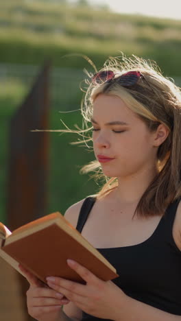 la mujer cierra el libro cubierto de marrón, tomando una respiración profunda mientras la luz del sol brilla sobre ella, el viento sopla suavemente su cabello, con una serena vegetación y estructura de madera en el fondo borroso