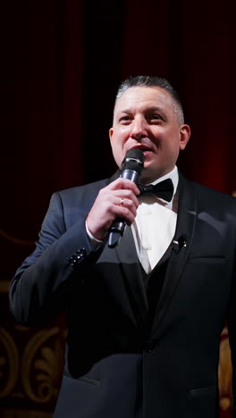 Male announcer with microphone on the stage. Actor in black suit on red scene curtains background. Vertical video