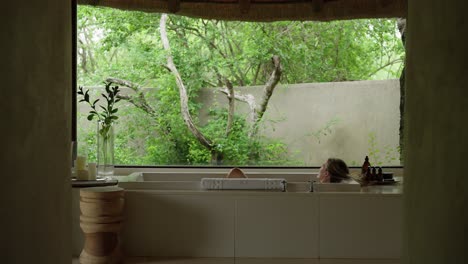 A-woman-lying-in-a-luxury-bath-with-a-large-window-looking-out-into-a-natural-landscape