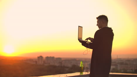 Un-Hombre-Con-Audífonos-En-El-Techo-Se-Relaja-Trabajando-Remotamente-Disfrutando-De-La-Vida-A-Pesar-De-Tomar-Una-Cerveza-Y-Escribir-En-El-Teclado.-Opere-En-La-Bolsa-De-Valores-Usando-Una-Computadora-Portátil-Y-Disfrutando-De-La-Hermosa-Vista