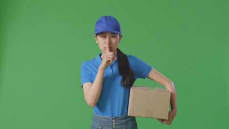 mensajera asiática en uniforme azul mostrando un gesto de silencio y sacudiendo la cabeza mientras entrega un cartón en el fondo de la pantalla verde en el estudio