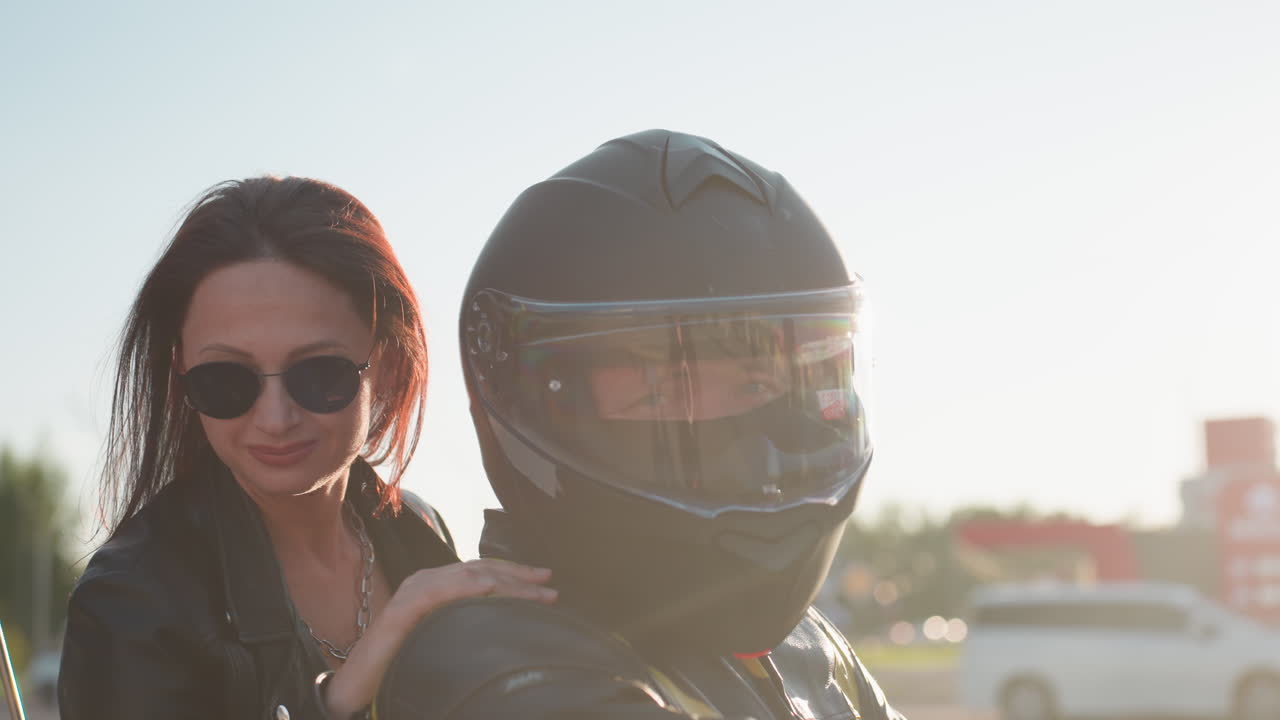 Couple on power bike look ahead calmly as sunlight creates glow around them, man wears helmet with covered visor while woman in sunglasses rests hand on his shoulder
