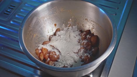 Pouring-flour-on-raw-seasoned-chicken-pieces-into-a-bowl,-close-up-view-in-home-kitchen