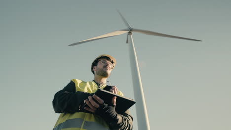 a professional engineer in a reflective vest and white security helmet uses a tablet to inspect wind turbines, emphasizing the role of renewable energy in protecting the environment