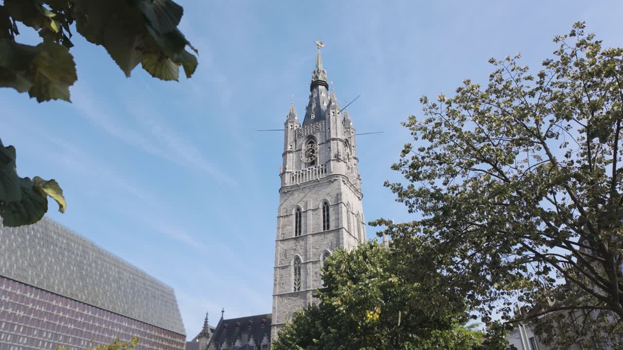 el campanario de gante, una histórica torre medieval en bélgica, se encuentra en medio de verdor verde bajo un cielo azul brillante, simbolizando una rica herencia cultural y esplendor arquitectónico.
