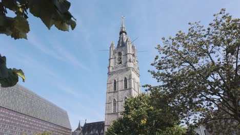 the belfry of ghent, a historic medieval tower in belgium, stands amidst verdant greenery under a bright blue sky, symbolizing a rich cultural heritage and architectural splendor.