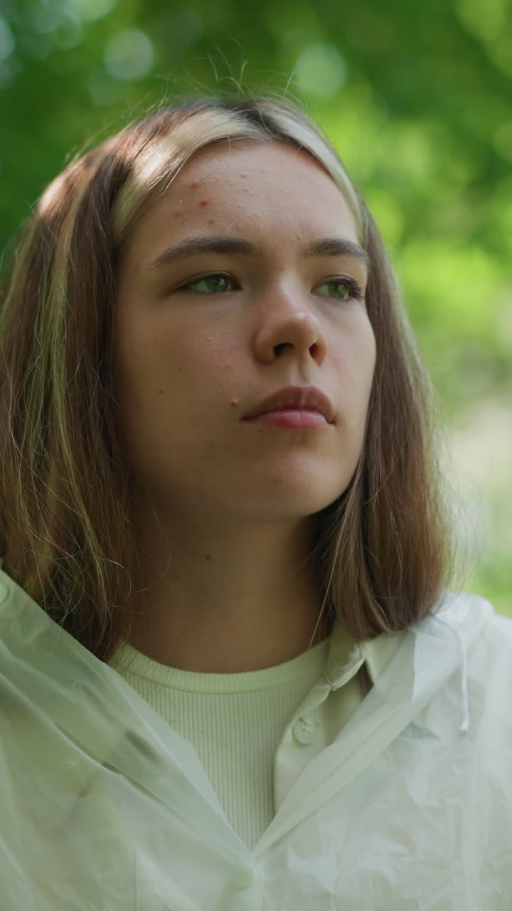 una joven con un impermeable translúcido está al aire libre, perdida en profundos pensamientos, con una expresión serena y reflexiva en su rostro, rodeada de árboles verdes y exuberantes, parece contemplativa.