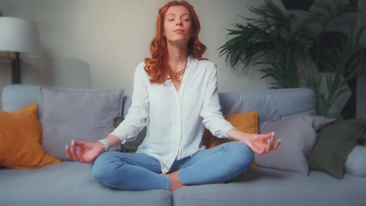 Redhead caucasian woman sits in lotus position on sofa doing yoga meditation