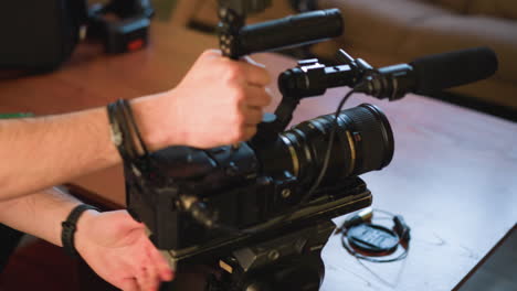 close-up of person adjusting camera on tripod with quick release plate. microphone attached to camera. green wall in background with soft indoor lighting, couch visible in distance