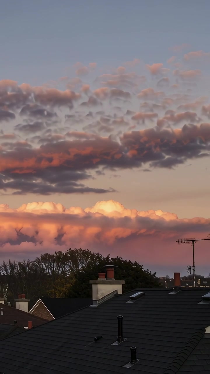 Vertical video: Drifting layered clouds changing color under sunset light over roofs, with antenna