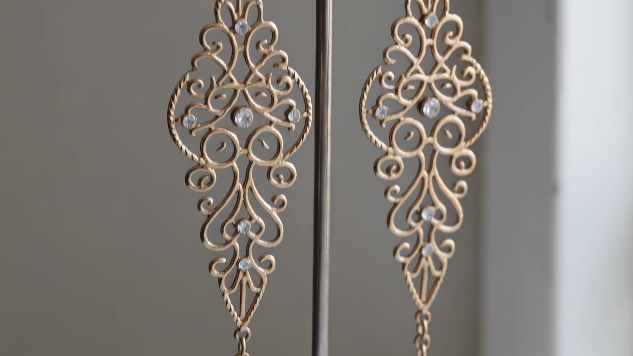 Câmera recuando, revelando um par de brincos pendentes de filigrana de ouro sobre a mesa, exibindo detalhes.