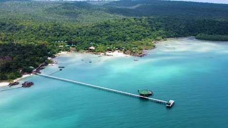 Aerial-establishing-shot-of-small-private-island-resort-surrounded-by-forrest-and-turquoise-blue-sea