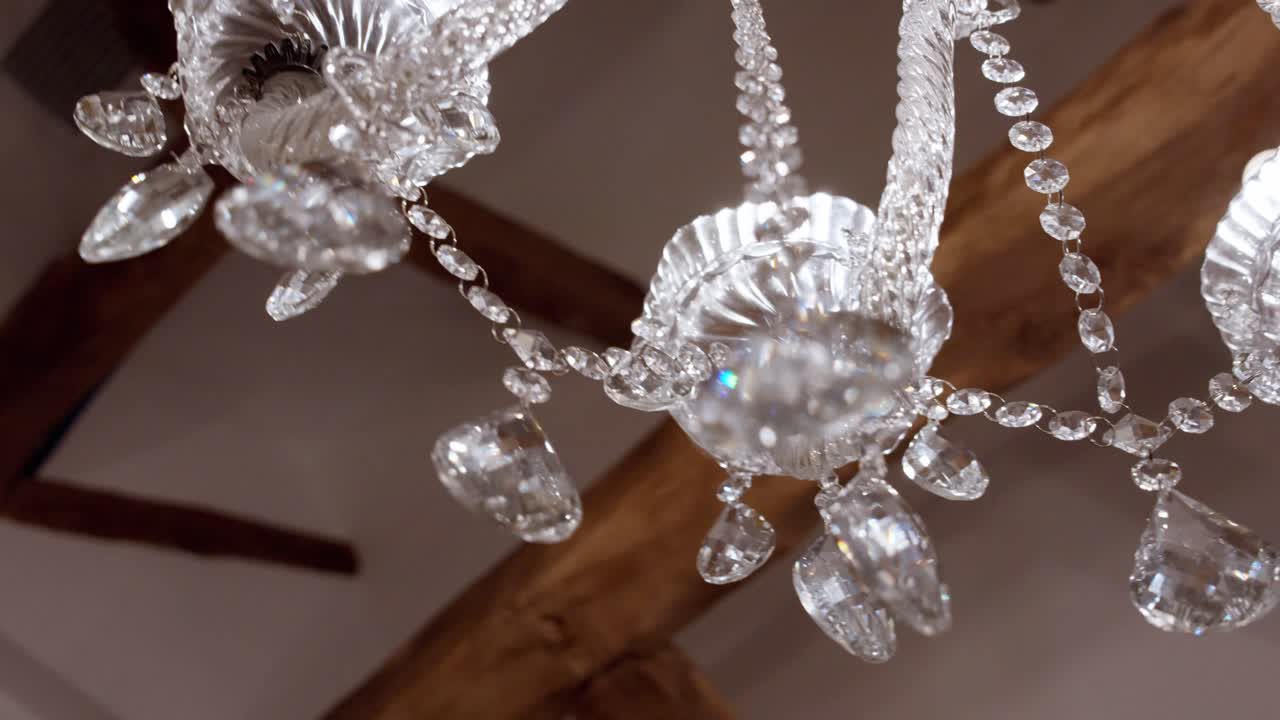 Lampadario di cristallo con pendenti e ornamenti in vetro trasparente, appeso a un soffitto interno con travi in legno che riflette la luce.