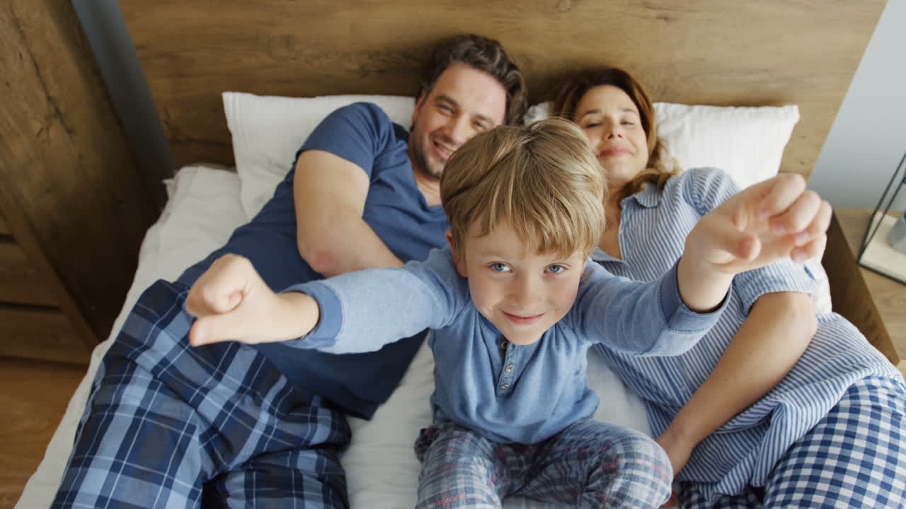 Top view of a little sitting on the bed and waving her hands to the camera between his parents who are lying