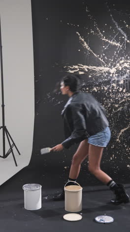 vertical video: young woman in leather jacket creating abstract art with paint splashes on wall