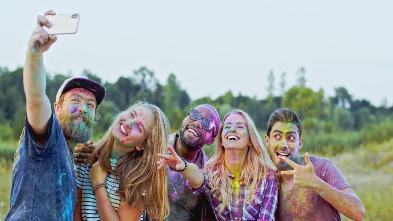 홀리 페스트 밖에서 셀카 사진을 찍는 동안 스마트폰 카메라에 미소를 짓는 화려한 페인트 가루를 입은 젊은 쾌활한 다민족 남성 및 여성 친구들