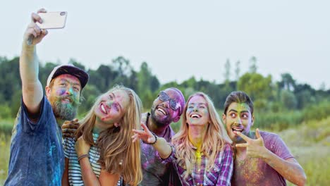 group of young cheerful multiethnic male and female friends in colorful paints powder smiling to the smartphone camera while taking selfie photo outsideat the holi fest