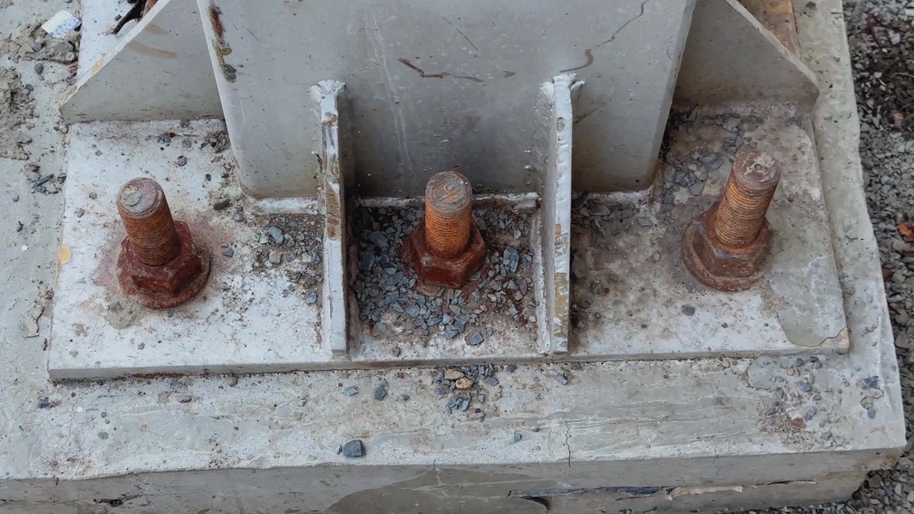 Ripresa ravvicinata in movimento di bulloni di ancoraggio in acciaio arrugginito fissati su una fondazione in cemento in un cantiere edile o infrastrutturale con dettagli strutturali industriali.