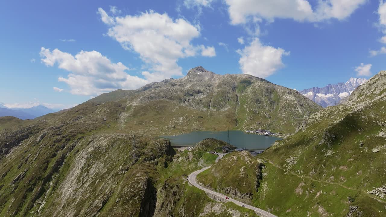 Vista aerea di una tortuosa strada di montagna che conduce a un lago alpino circondato da cime aspre. Le auto sono visibili sulla strada serpeggiante, enfatizzando il viaggio e l'esplorazione nelle Alpi svizzere