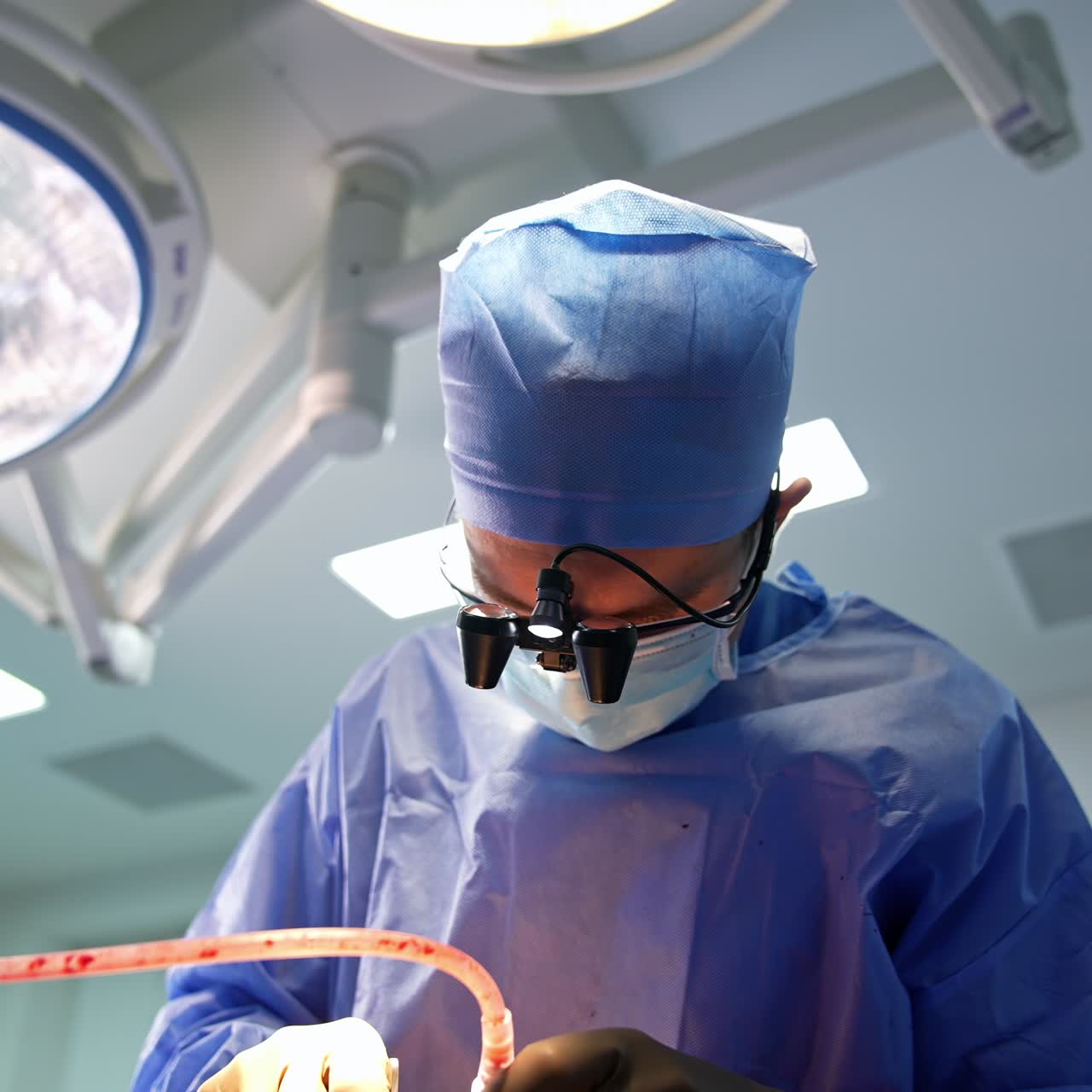 Overkrop af en mandlig læge, der er bøjet over patienten. Kirurgen bruger omhyggeligt et instrument med beskyttelsesbriller på. Lav vinkel.