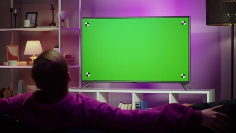 hombre viendo la televisión en la sala de estar por la noche. joven sentado en el sofá contra la televisión con pantalla verde croma, vista trasera. persona masculina cambiando canales usando el control remoto, relajándose.