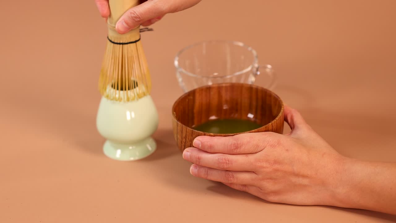 Hands whisk matcha green tea in wooden bowl using bamboo whisk, minimal background, soft lighting