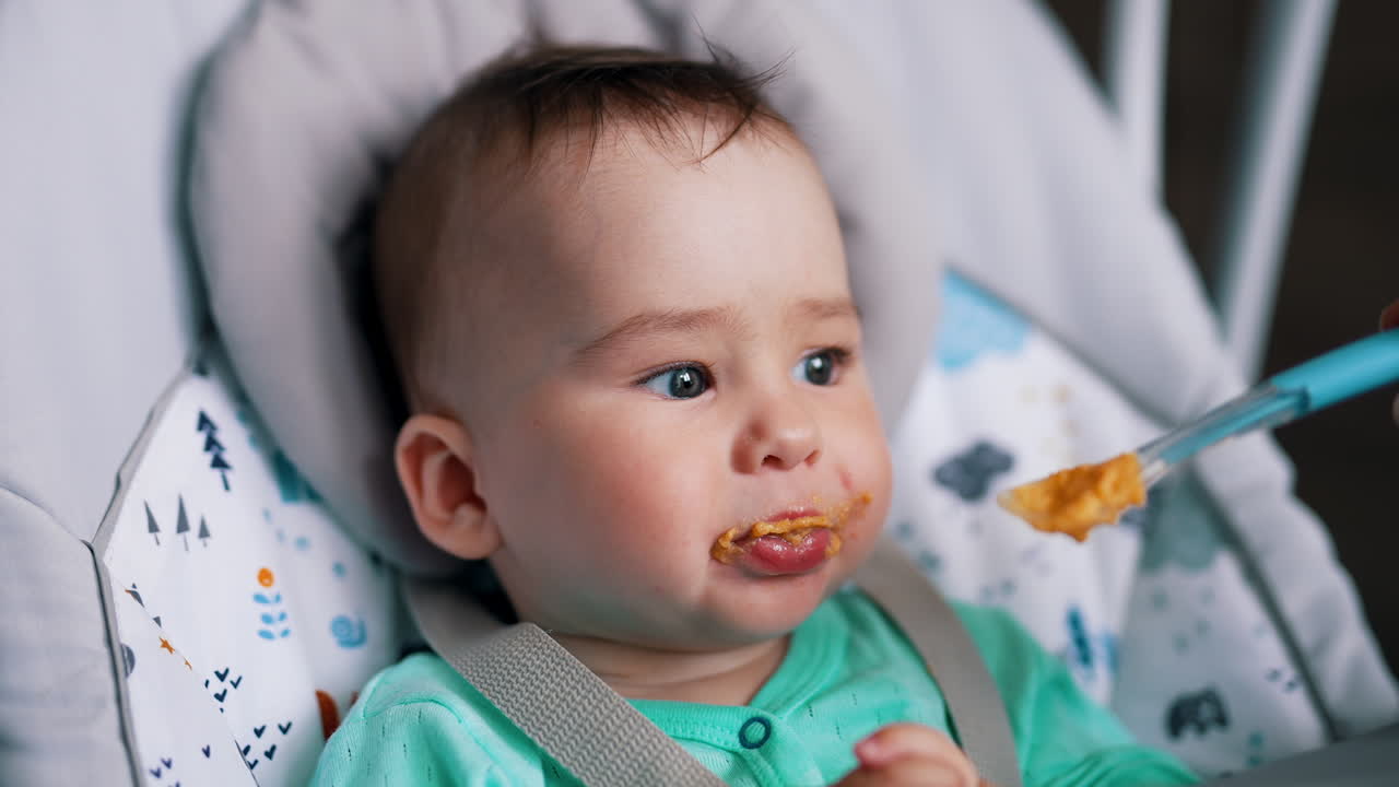 Lovely kid being fed from a special baby spoon. Beautiful toddler sitting in chair having meals. Close up.