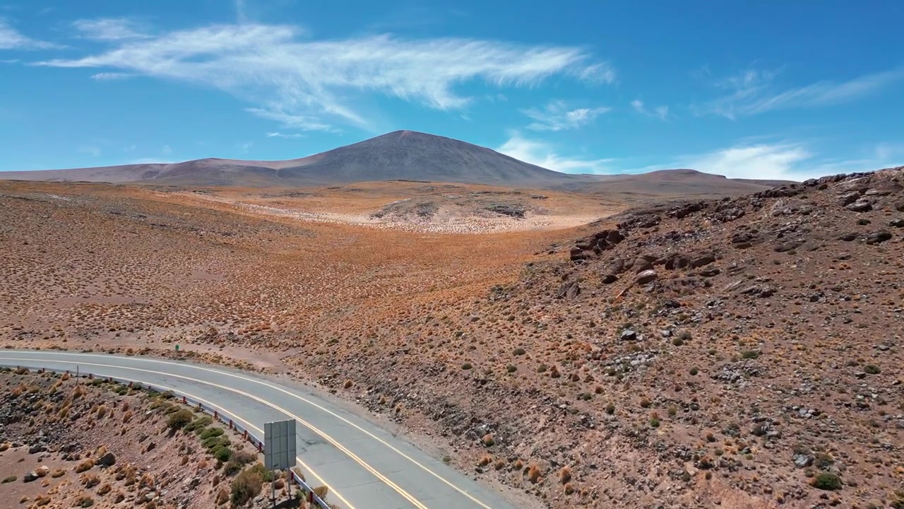 Volo in avanti su una strada di montagna tortuosa che attraversa una valle desertica andina rocciosa verso un ampio cono vulcanico sotto un cielo azzurro vivido