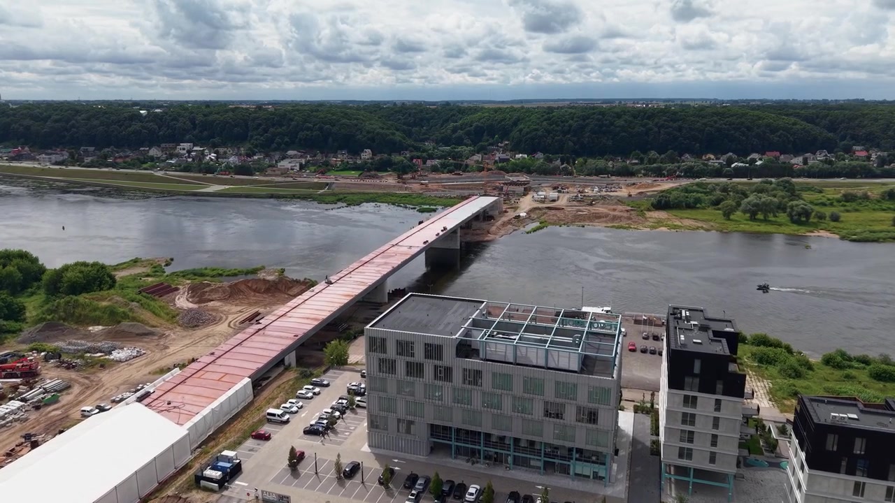 Dynamic aerial footage showcases a partially completed bridge spanning a wide river, modern office buildings nearby, lush landscape, overcast sky, and steady drone camera movement.