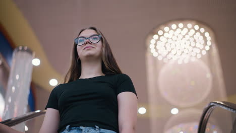 dama de tapa negra descendiendo escalera mecánica con postura elegante, fondo con luces bokeh y candelabro en un centro comercial moderno, iluminación suave y cálida y reflejos