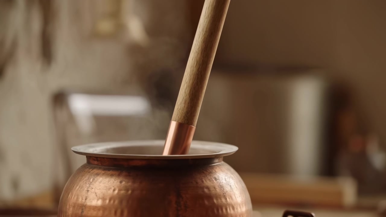 Close view of wooden pestle with rose-gold metal ferrule resting inside wide hammered copper mortar, tip touching inner rim while faint steam rises above vessel in warm rustic kitchen light