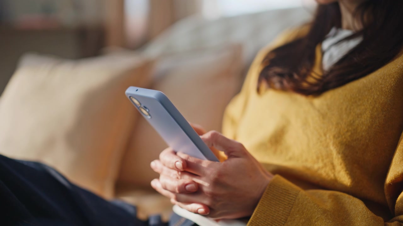Primo piano delle mani di una ragazza che scrive messaggi al cellulare, rilassandosi nel salotto di casa.
