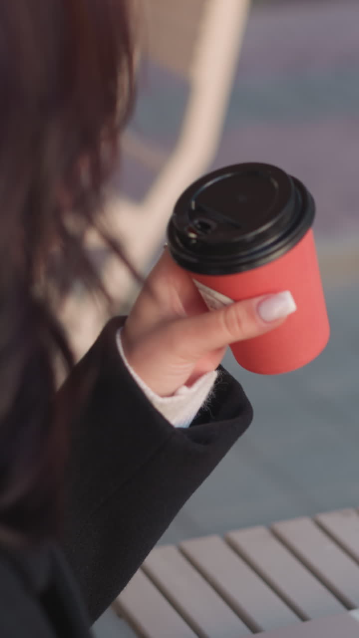 Close-up of student in black coat sipping red coffee cup outdoors while typing on laptop, setting cup down with focus on work, background features a chair and urban setting