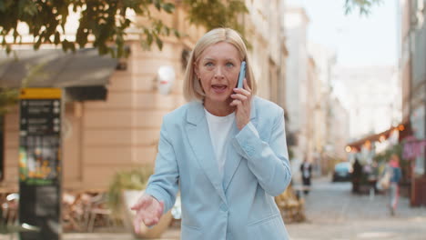 angry mature caucasian businesswoman arguing while talking on smartphone standing on city street