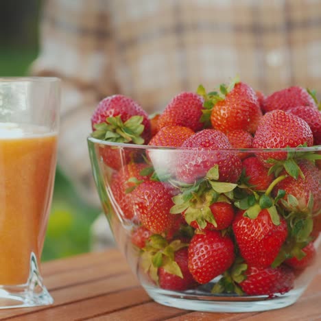 Der-Bauer-Stellt-Auf-Den-Tisch-Eine-Transparente-Schüssel-Mit-Frischen-Erdbeeren-Neben-Einem-Glas-Saft