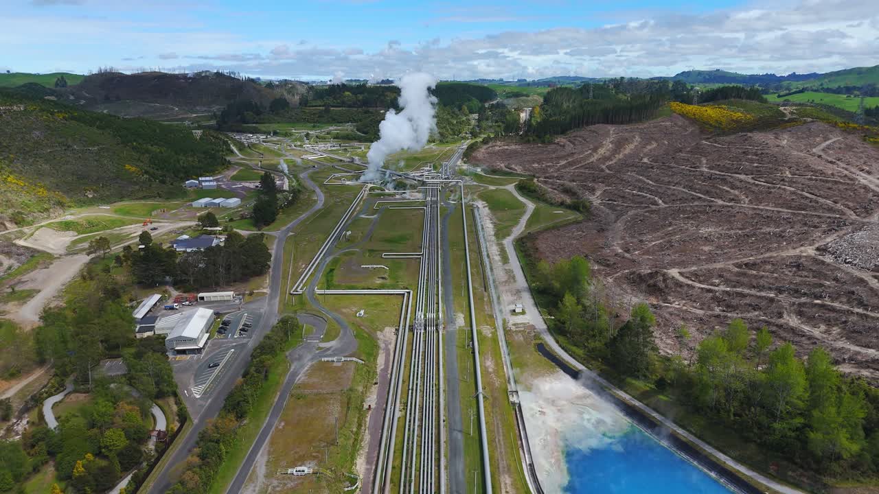 Tuberías de una planta geotérmica que conducen a vapor ascendente junto a un estanque azul brillante cerca de Taupo, Nueva Zelanda, en una toma aérea amplia de dron frontal.
