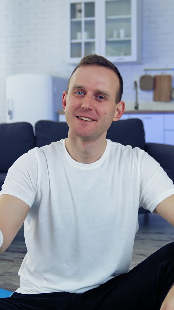 Man with a bottle of water at home. Young man in white t-shirt sitting on a floor and showing a bottle after doing fitness exercises. Vertical video