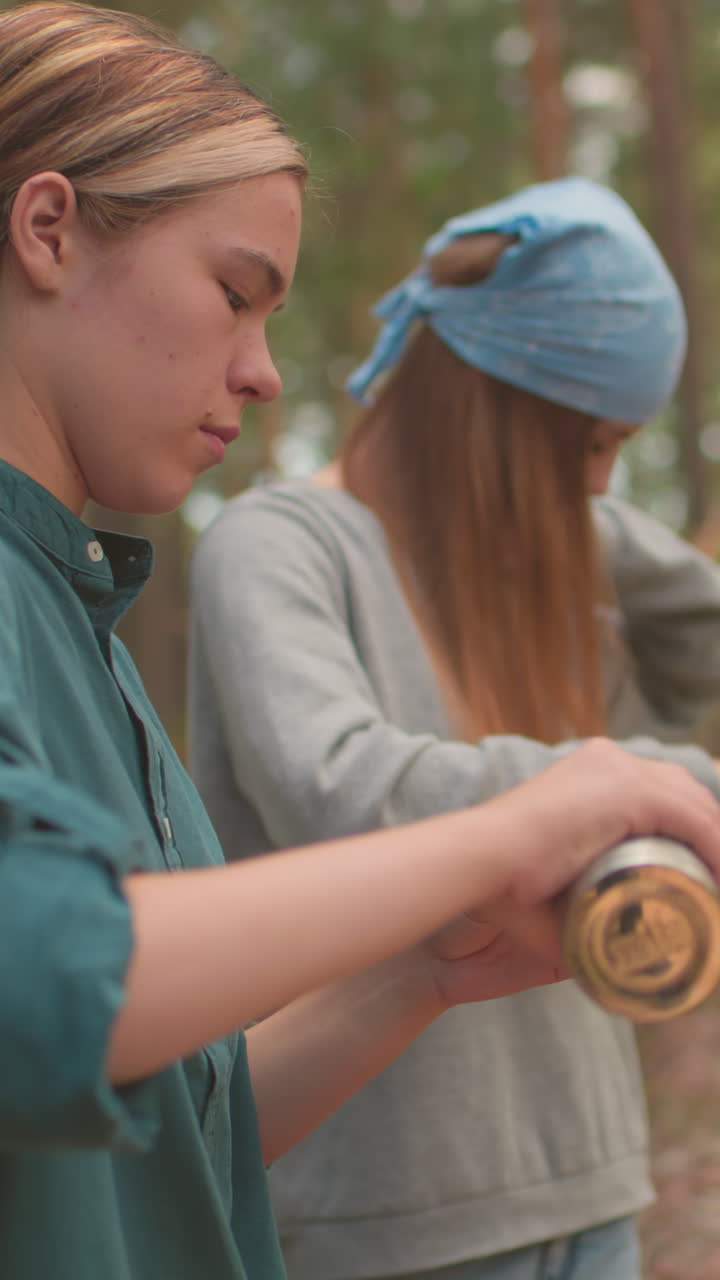 пешеходы делают спокойный перерыв в пышном лесу, один осторожно открывает термос, в то время как другой осторожно чистит волосы, рюкзаки отдыхают на упавшем дереве, захватывая расслабленный, спокойный момент, когда они наслаждаются природой