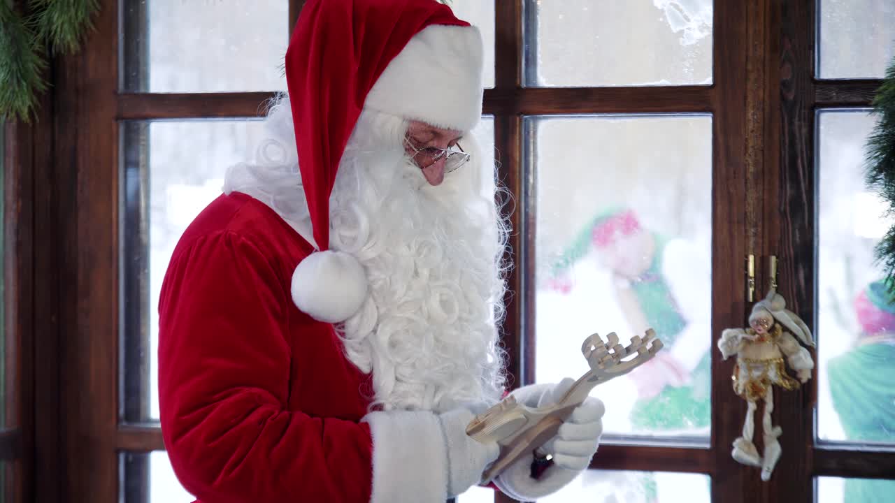 Santa Claus in red costume with wooden toy indoors. Santa shows hand gesture through the window to elves playing in snow