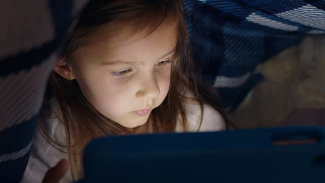 niña linda usando una tableta digital bajo una manta disfrutando de aprender en la tecnología de pantalla táctil jugando juegos divirtiéndose antes de acostarse